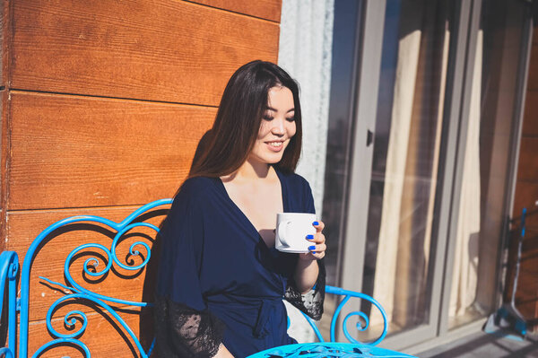 Beautiful Asian smiling woman enjoying coffee or tea on sunny balcony. Young happy lady in blue bathrobe chilling sunbathing on terrace at summertime. Royalty Free Stock Photos