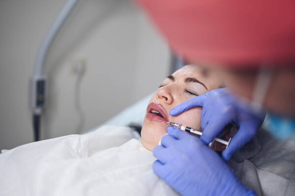 Cosmetologist doing painful lip augmentation procedure with hyaluronic acid. The beautician pierces lips by needle. Woman suffering subcutaneous injection to increase lips shape with dermal filler Stock Image