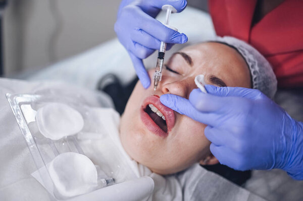 Cosmetologist doing painful lip augmentation procedure with hyaluronic acid. The beautician pierces lips by needle. Woman suffering subcutaneous injection to increase lips shape with dermal filler Royalty Free Stock Images