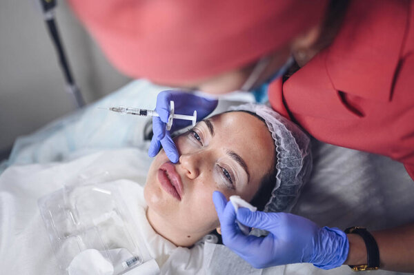 Cosmetologist doing painful lip augmentation procedure with hyaluronic acid. The beautician pierces lips by needle. Woman suffering subcutaneous injection to increase lips shape with dermal filler Royalty Free Stock Photos