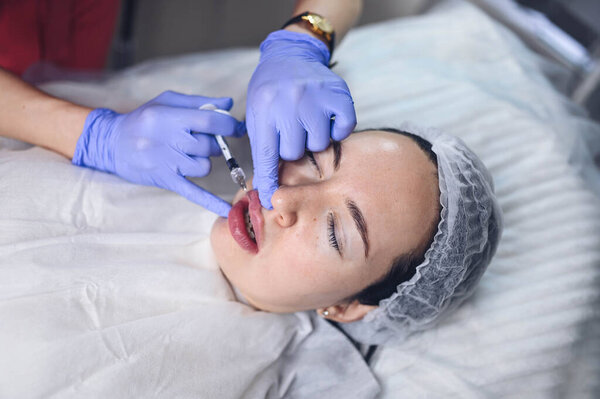 Cosmetologist doing painful lip augmentation procedure with hyaluronic acid. The beautician pierces lips by needle. Woman suffering subcutaneous injection to increase lips shape with dermal filler Stock Picture