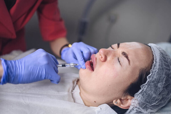 Cosmetologist doing painful lip augmentation procedure with hyaluronic acid. The beautician pierces lips by needle. Woman suffering subcutaneous injection to increase lips shape with dermal filler Royalty Free Stock Photos