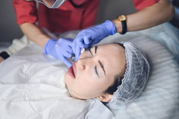 Cosmetologist doing painful lip augmentation procedure with hyaluronic acid. The beautician pierces lips by needle. Woman suffering subcutaneous injection to increase lips shape with dermal filler Stock Photo