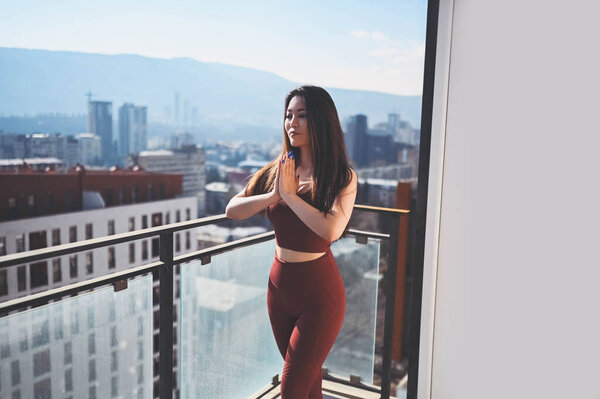 Beautiful Asian smiling fit woman doing yoga on sunny balcony in sport suit. Young happy lady on terrace at summertime. Big city view. Sports exercises and gymnastics, healthy way of life concept. Stock Image