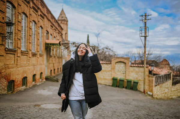 Young happy smiling woman tourist traveling alone walking with attractions in an empty street in small old town in Europe. Brunette lady in black down jacket and white hoodie Stock Image