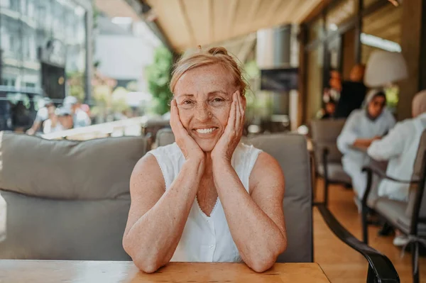 Happy senior woman with perfect smile and teeth sitting outdoors at summer sidewalk street cafe or restaurant terrace. Retired mature people holiday vacation, active lifestyle concept