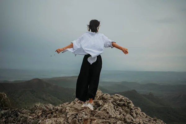 Happy woman tourist hiking up mountain enjoying nature. Landscape in cloudy gloomy windy weather. Female in white shirt with open arms outstretched in joy, enjoying travel. Freedom concept