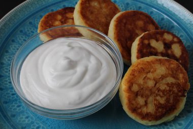 Kahvaltı peynirli kekler birkaç parça krep beyaz soslu ekşi kremalı büyük mavi bir tabak üzerinde yatıyor. Siyah arka planda geleneksel Rus Slav kahvaltısı..