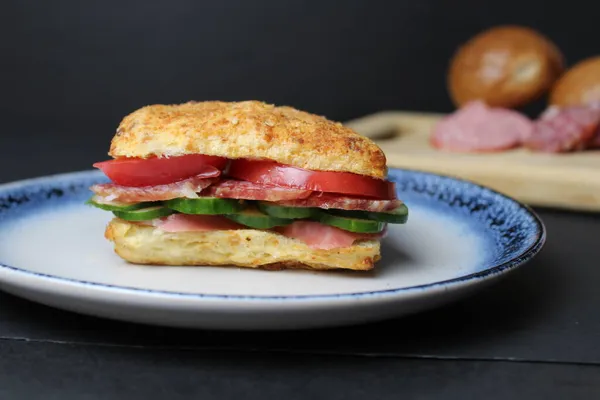 sandwich sandwich of white square bun with vegetables tomato cucumber sausage ham on a blue plate. in the background is a tray board with ingredients sausage vegetables tomatoes on a black background. Lunch snack delicious homemade.