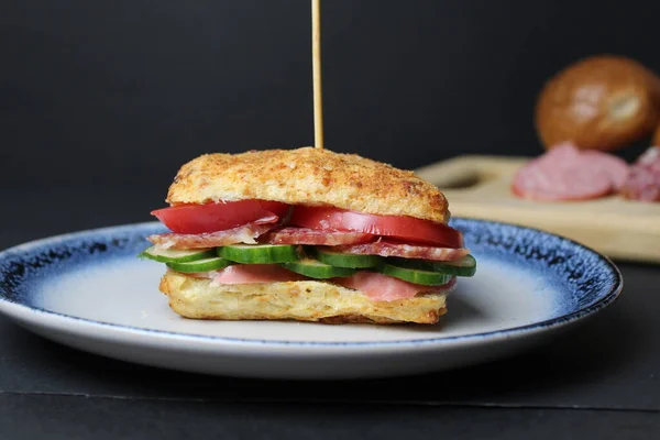 sandwich sandwich of white square bun with vegetables tomato cucumber sausage ham on a blue plate. in the background is a tray board with ingredients sausage vegetables tomatoes on a black background. Lunch snack delicious homemade.