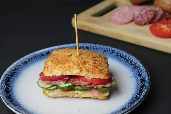 sandwich sandwich of white square bun with vegetables tomato cucumber sausage ham on a blue plate. in the background is a tray board with ingredients sausage vegetables tomatoes on a black background. Lunch snack delicious homemade.