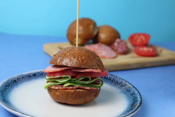 sandwich, burger with buckwheat brown bun vegetables pieces of tomatoes cucumbers on a blue plate on a blue background are the ingredients for making a burger vegetables sausage ham.