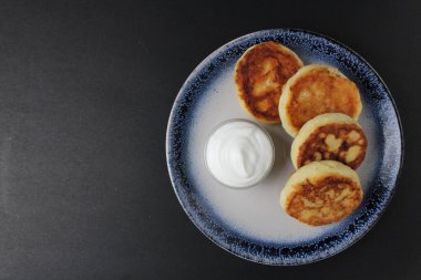 Üç cheesecake mavi bir tabağın üzerinde, siyah bir zemin üzerinde beyaz ekşi krema sosu ve yazı için yeri olan bir sos kasesi üzerinde uzanıyor. Rus kahvaltısı gelenekseldir. Beslenme düzenine uygun. süzme peynir yemekleri..