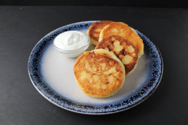 Üç cheesecake mavi bir tabağın üzerinde, siyah bir zemin üzerinde beyaz ekşi krema sosu ve yazı için yeri olan bir sos kasesi üzerinde uzanıyor. Rus kahvaltısı gelenekseldir. Beslenme düzenine uygun. süzme peynir yemekleri..