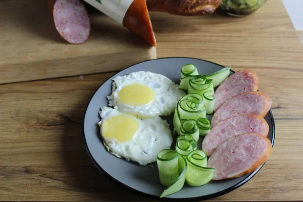 朝食の伝統的なスクランブルエッグ2ハムソーセージとキュウリサラダテーブルの上の灰色のプレートに 隣には朝食ソーセージパンを作るための商品があります 横写真 — ストック写真