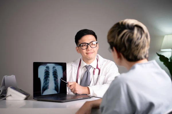 Kind and polite young Asian medical doctor orthopedic surgeon wearing stethoscope showing x-ray image from laptop while explaining and soothing patient about diagnosis and treatment plan in examination room in hospital. Asian qualified physician usin