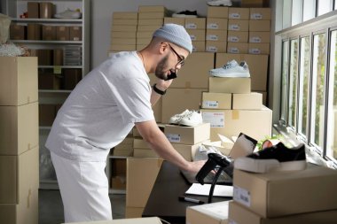 Asyalı adam online satıcı, telefondaki ve dizüstü bilgisayardaki siparişleri onaylıyor. E-ticaret erkek işyeri sahibi telefon kullanıyor ve depodaki dizüstü bilgisayara bakıyor..