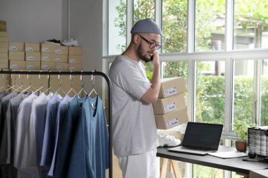 Asyalı adam online satıcı, telefondaki ve dizüstü bilgisayardaki siparişleri onaylıyor. E-ticaret erkek işyeri sahibi telefon kullanıyor ve ambarda paketler tutuyor..