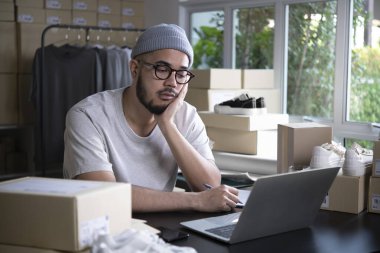 Çevrimiçi satıcı girişimci bir depoda dizüstü bilgisayara bakıyor ve satış ve müşteri eksikliği nedeniyle e-ticaret işi hakkında endişeleniyor.
