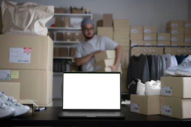 Depodaki masaüstünde duran boş beyaz ekranlı dizüstü bilgisayar. Asyalı adam müşteriye göndermeden önce hisse senedi ve envanter kontrolü yapıyor. Mağaza deposunda çalışan bir e-ticaret şirketi sahibi..