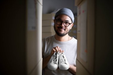 Başarılı Asyalı genç adam perakende satıcısı, girişimci, çevrimiçi mağaza teslimatçısı küçük işletme sahibi ürünleri gösteriyor ve teslimat deposunda paket kutularıyla kameraya bakıyor..