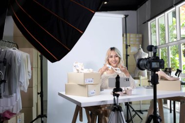 Genç, çekici Asyalı kadın blogcu ya da video blogcusu ürünü inceleyen kameraya bakıyor. Modern iş kadını pazarlama için sosyal medyayı kullanıyor. Sosyal medya kavramı üzerinde iş çevrimiçi etkileyiciName.