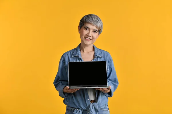Portrait of middle aged Asian woman 50s wearing casual denim shirt white t-shirt holding laptop computer isolated on yellow background, looking and smiling at the camera.