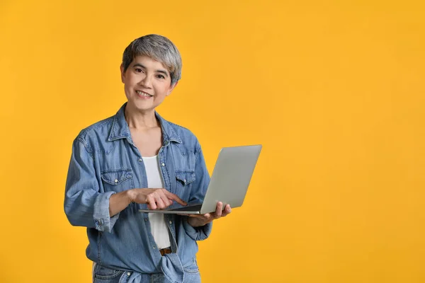 Portrait of middle aged Asian woman 50s wearing casual denim shirt white t-shirt holding laptop computer and pointing fingers isolated on colour background, looking and smiling at camera