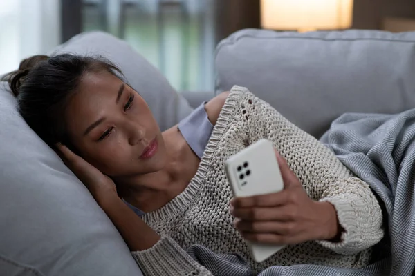 Frustrated and depressed young Asian woman is crying with a smartphone in hand while she lying in the couch at home, sad Asian female on sofa feel down read bad news on cellphone online.