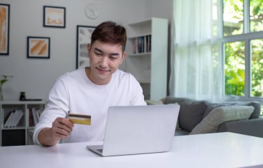 Kredi kartı tutan ve dizüstü bilgisayar kullanan genç bir Asyalı. Evde çalışan bir iş adamı. Çevrimiçi alışveriş, e-ticaret, internet bankacılığı, harcama, ev konseptinden çalışma