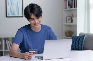 Kredi kartı tutan ve dizüstü bilgisayar kullanan genç bir Asyalı. Evde çalışan bir iş adamı. Çevrimiçi alışveriş, e-ticaret, internet bankacılığı, harcama, ev konseptinden çalışma