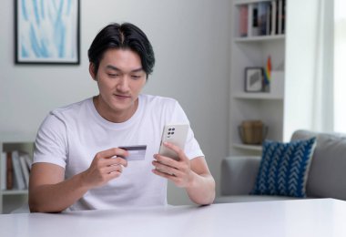 Genç Asyalı adam kredi kartı tutuyor ve akıllı telefon kullanıyor. Evde çalışan bir iş adamı. Çevrimiçi alışveriş, e-ticaret, internet bankacılığı, harcama, ev konseptinden çalışma