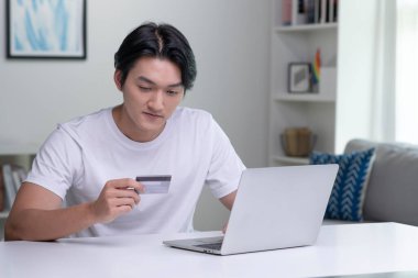 Kredi kartı tutan ve dizüstü bilgisayar kullanan genç bir Asyalı. Evde çalışan bir iş adamı. Çevrimiçi alışveriş, e-ticaret, internet bankacılığı, harcama, ev konseptinden çalışma