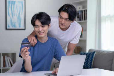 Kredi kartı tutan ve dizüstü bilgisayar kullanan genç Asyalı eşcinsel çift. LGBT gay iş adamı evde çalışıyor. Çevrimiçi alışveriş, e-ticaret, internet bankacılığı, harcama, ev konseptinden çalışma