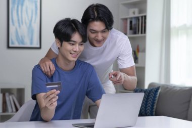 Kredi kartı tutan ve dizüstü bilgisayar kullanan genç Asyalı eşcinsel çift. LGBT gay iş adamı evde çalışıyor. Çevrimiçi alışveriş, e-ticaret, internet bankacılığı, harcama, ev konseptinden çalışma