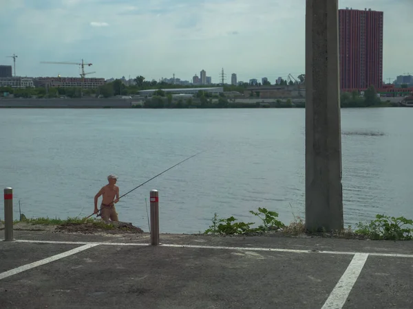 An old man in shorts and a cap and a fishing rode in his hands pulling fish out of the Dnipro river on an urban beach near the North bridge in Kyiv city, Ukraine. Active hobby concept.