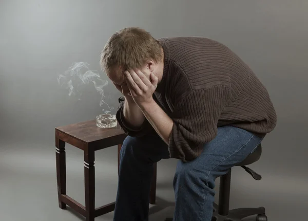 Depressed young man sitting on a chair, smoking holding his head — Stock Photo, Image