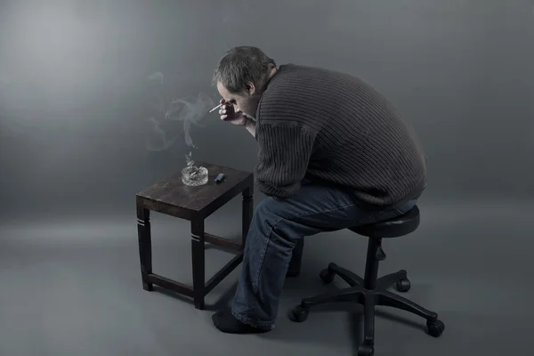 Depressziós fiatal férfi ült egy széken, cigarettázott Stock Fotó