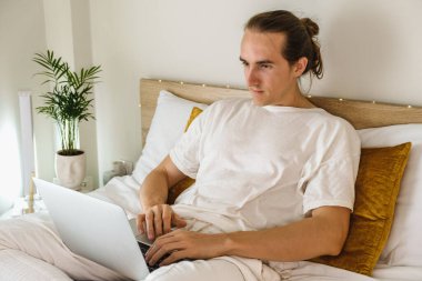 Evde yatakta oturan ve dizüstü bilgisayarda yazan yakışıklı genç adam.