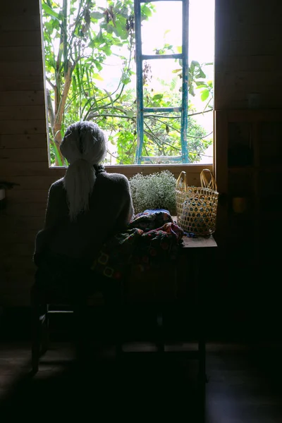 Old woman sitting near window with handmade product, yarn colorful blanket by crochet from yarn with old style, window light make shadow on wooden table of classic style