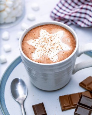 Kar tanesi şeklinde kremalı ev yapımı sıcak çikolata ve beyaz bardakta tıraş edilmiş çikolata. Marşmelovlar arka plana saçılmış..