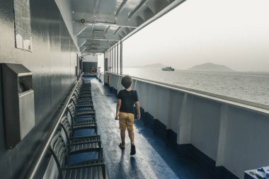Çocuk feribotta sandalyelerin yanında tek başına yürürken güzel denize bakıyor.
