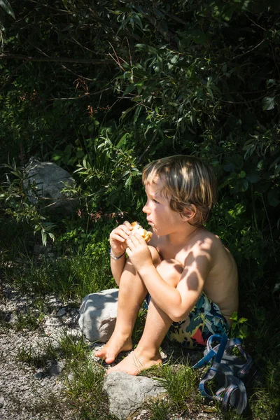 Menino Comendo Hambúrguer Enquanto Sentado Grama Sob Pequenos Arbustos Dia — Fotografia de Stock