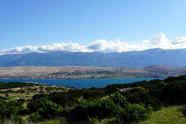 Büyük yeşil dağın ve mavi denizin üzerindeki mavi gökyüzünde beyaz bulutlar ve önplandaki yeşil bitki örtüsü.