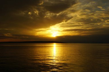 Gökyüzünde koyu ağır bulutlar ve gün batımında altın deniz manzarasının arka planında güneş.