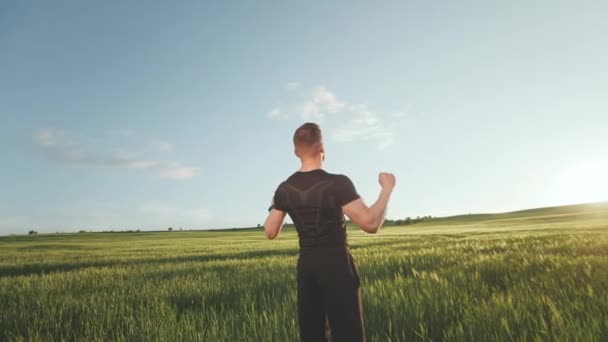 Un hombre con uniforme deportivo está parado en medio de un campo. Está mirando alegremente al atardecer y levantando las manos en el aire. 4K — Vídeo de stock