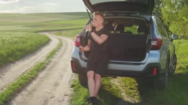 Spor üniformalı genç bir adam bir arabanın bagajında oturuyor. Su içiyor ve etrafındaki doğaya bakıyor. O, elinde bir akıllı telefon tutuyor. Güneş ışıl ışıl parlıyor. 4K
