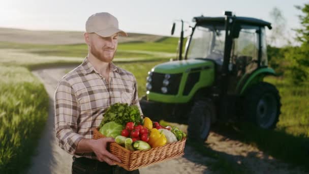 Um agricultor está de pé perto de um campo e segurando um cesto de legumes do campo. Ele está levantando a cabeça e olhando para a câmera. Há um tractor no fundo. 4K — Vídeo de Stock