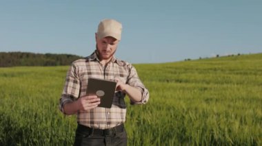 Çiftçi tarlanın yanında duruyor ve bir tabletle çalışıyor. Başını kaldırıyor ve kameraya bakıyor. Kafasında bir kep var. Alan arka planda. 4K