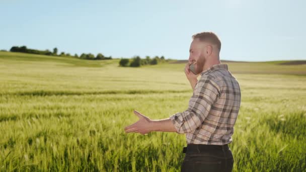 Un joven granjero está parado junto al campo y hablando emocionalmente por teléfono. Está saludando y sonriendo. El campo está en el fondo. 4K — Vídeos de Stock